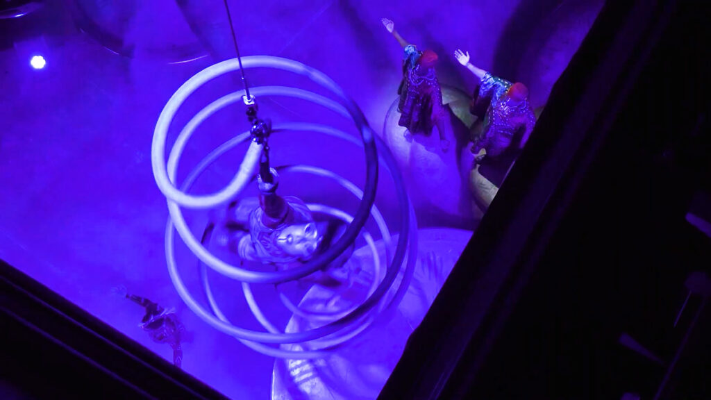 Vue plongeante depuis la régie sur la scène de l'Opéra, baignée de lumière violette. Une structure en spirale blanche descend du plafond, tandis que des artistes en costumes colorés effectuent une performance en dessous.