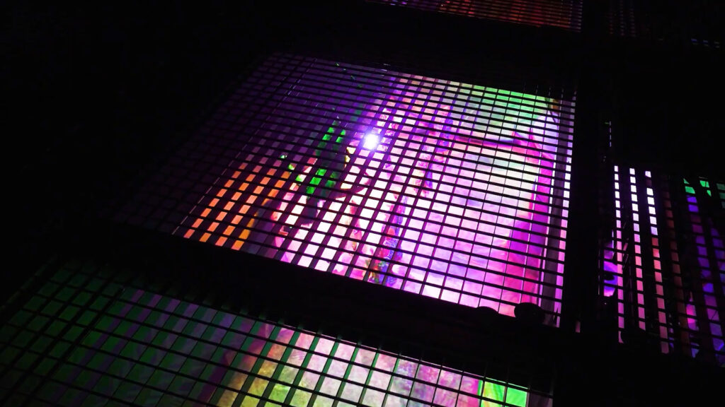 Vue à travers une grille métallique du plafond technique de l'Opéra. En contrebas, la scène est baignée de lumières roses, violettes et vertes. On y aperçoit des danseurs en costumes colorés en pleine performance, vus de très haut.