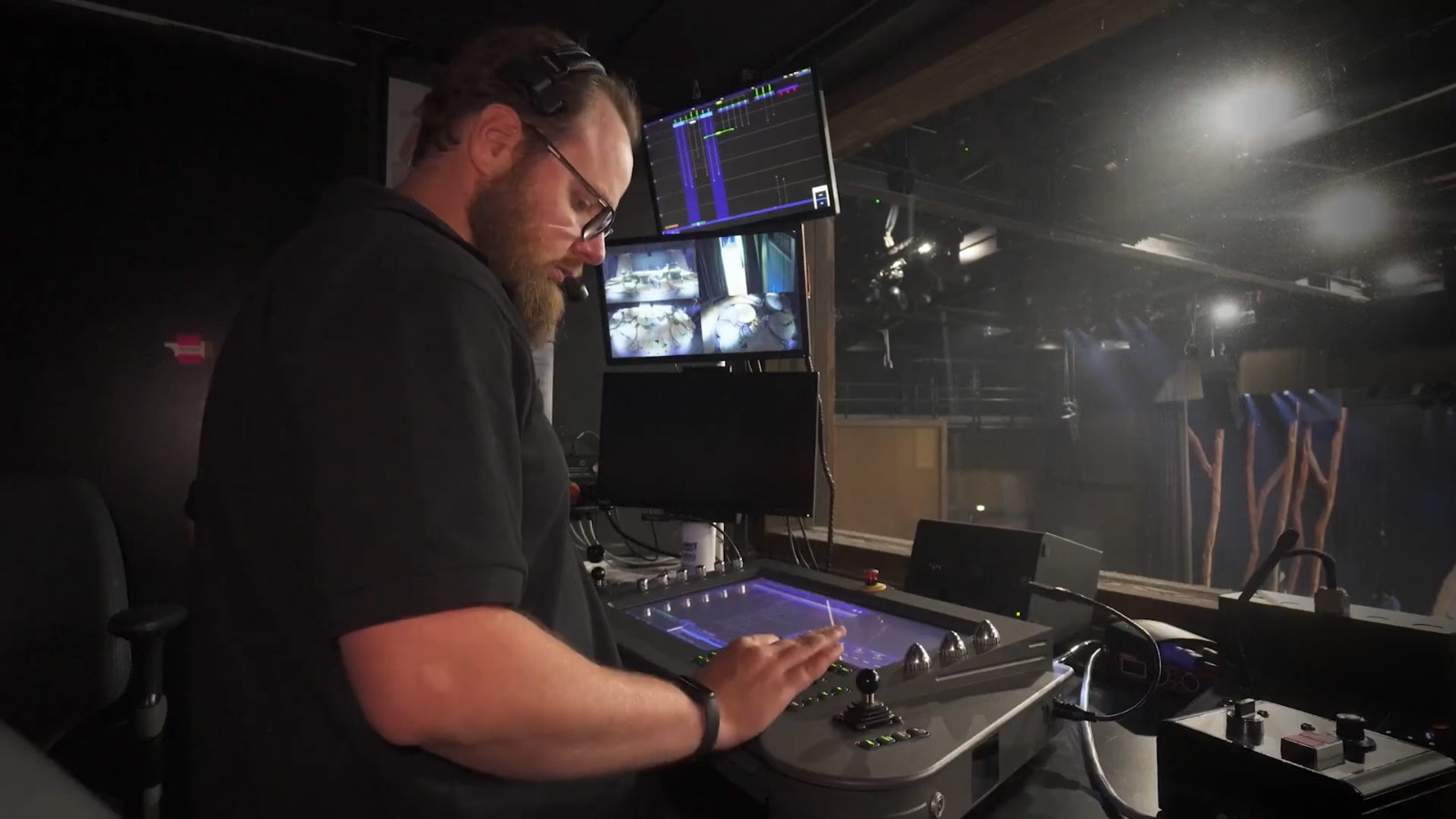 Gros plan sur un stage manager à son poste de travail dans les coulisses de l'Opéra Bastille. Il porte un casque de communication et manipule avec précision une console technique, les yeux fixés sur ses écrans de contrôle.