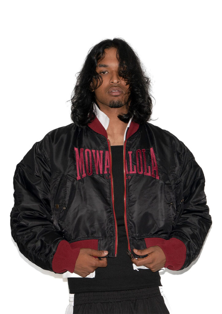 Un homme aux longs cheveux noirs ondulés et à la barbe courte pose devant un fond blanc uni. Il porte une veste de type bomber noire et volumineuse avec des poignets et un col bordeaux. Le nom « MOWALOLA » est brodé en grandes lettres bordeaux sur le devant de la veste. Il porte également un haut noir et un pantalon de sport assorti avec une bande latérale blanche.