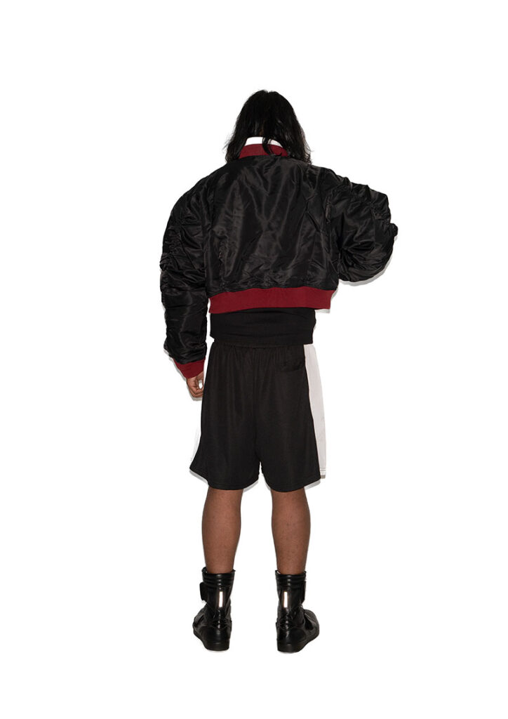 Vue de dos d'un homme en bomber noir court à col et taille bordeaux, short de sport noir à bandes blanches et bottes montantes noires sur fond blanc.